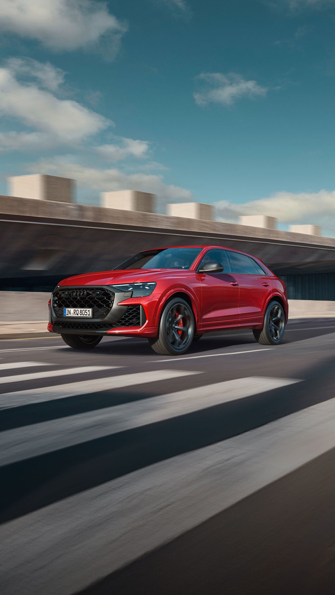 A red Audi Q8 SUV driving on a city street, with modern buildings and a blue sky in the background.