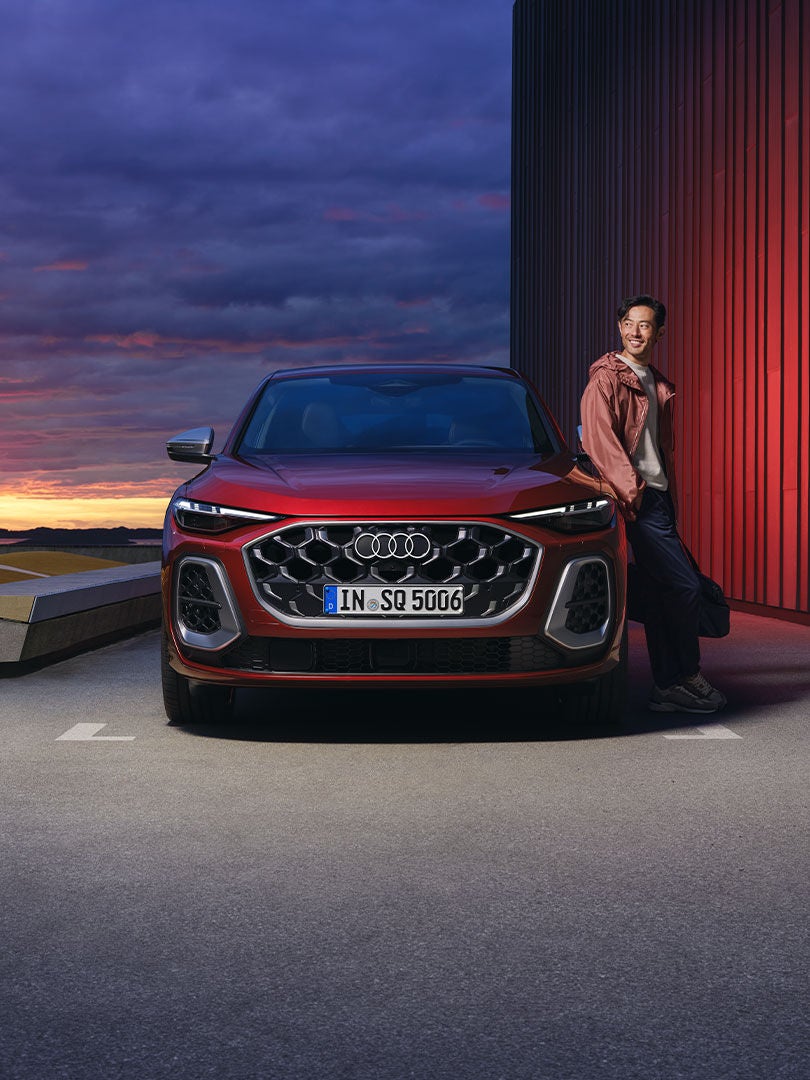 Der rote SQ5 Sportback steht auf einem Parkplatz parallel zu einem Hafen. Ein junger Mann lehnt sich an das Fahrzeug.