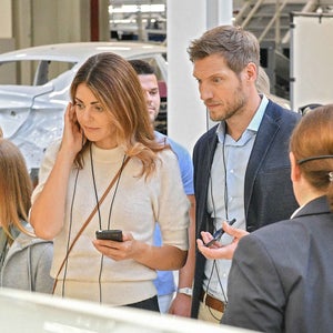 Frau und Mann mit Audioguide hören einer Führung zu; die Frau hält ein Smartphone, weitere Personen im Hintergrund.