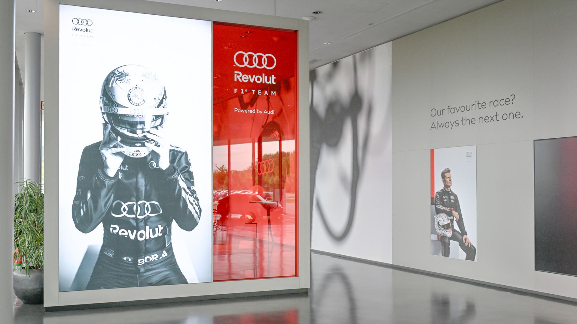 Heller Flur mit großem Foto eines Audi-Rennfahrers, rotem Revolut-F1-Team-Schaukasten und einem weiteren Rennfahrerporträt an der Wand.