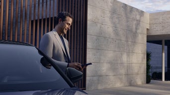 Person hält Smartphone mit Fahrzeug-App in der Hand, im Hintergrund ein geparkter weißer Audi.