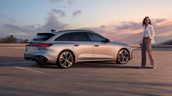 Eine Frau steht neben einem silbernen Audi A5 Avant auf einem offenen Parkplatz bei Sonnenuntergang.