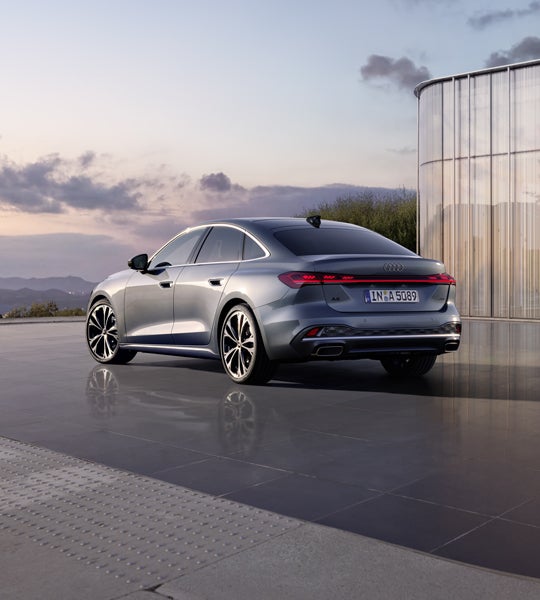 Die A5 Limousine im schrägen Heckprofil. Das Fahrzeug spiegelt sich auf dem glatten Untergrund. Im Hintergrund sind die Konturen von Bergen bei Dämmerung zu sehen.