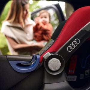 A child car seat with the Audi logo on it with A mum holding a baby in the background