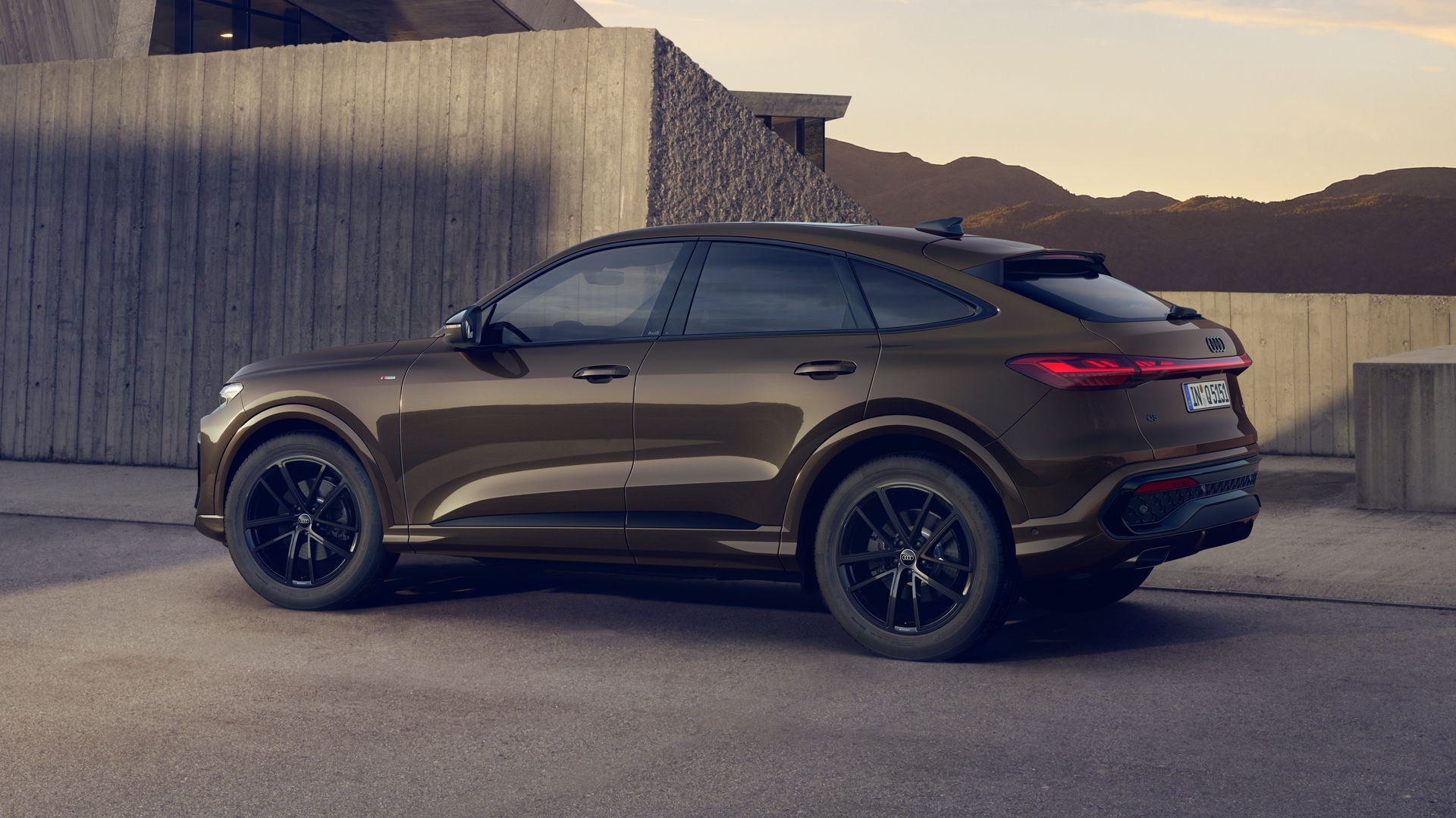 Der Q5 Sportback mit der Individuallackierung Teakbraun Metallic steht im Seitenprofil vor einem Gebäude. Im Hintergrund sind Berge zu erkennen.