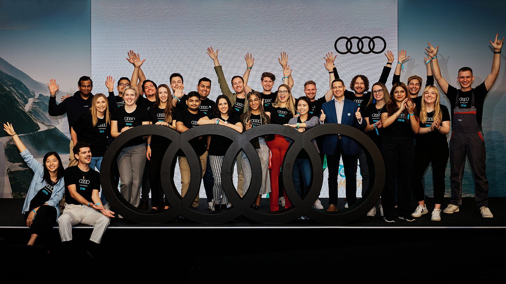 A group of people raise their arms and stand in front of a large black Audi logo