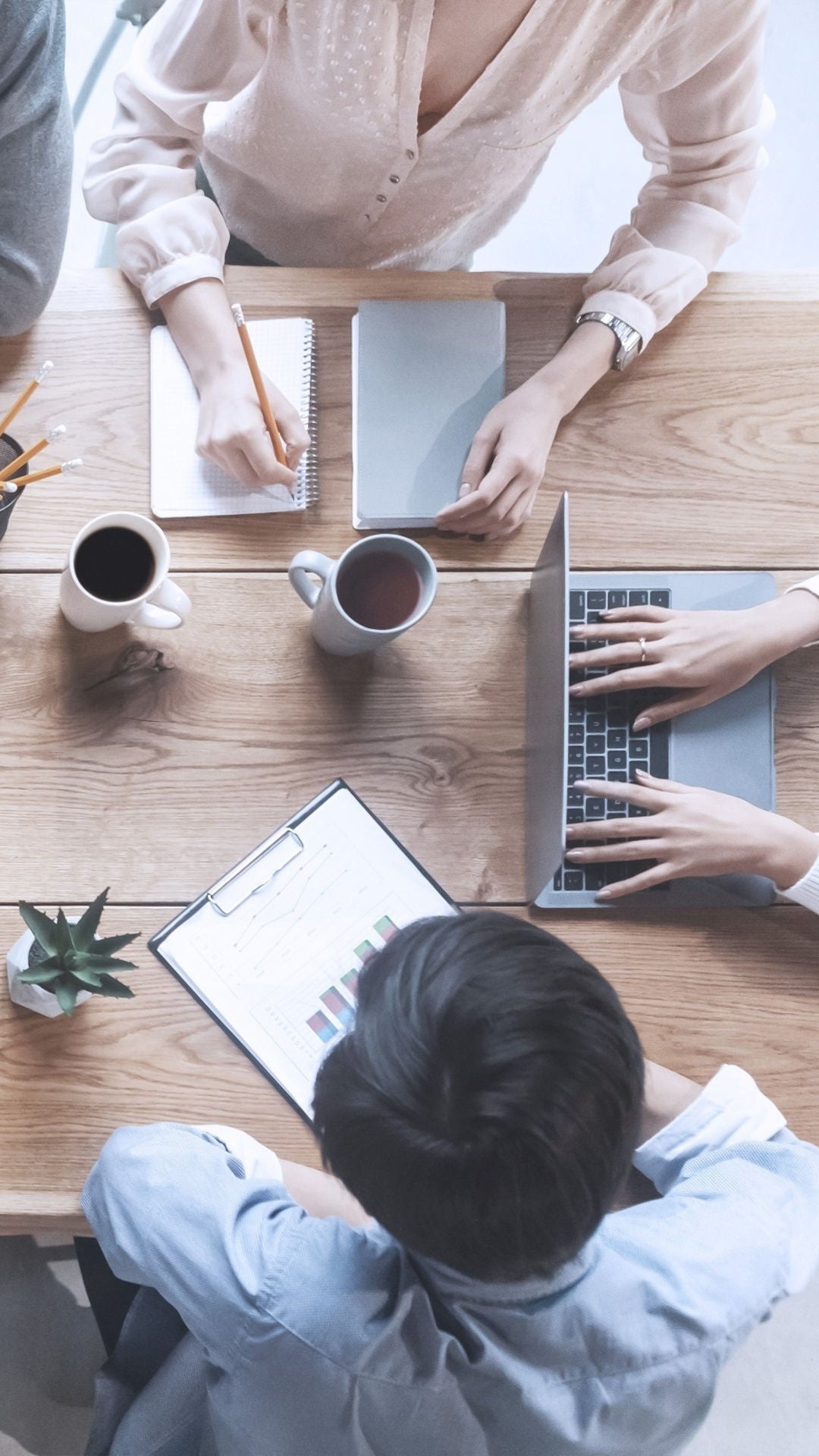 A number of people are sitting at a table and working together. Laptops, tablets, notes and coffee cups can be seen on the table.