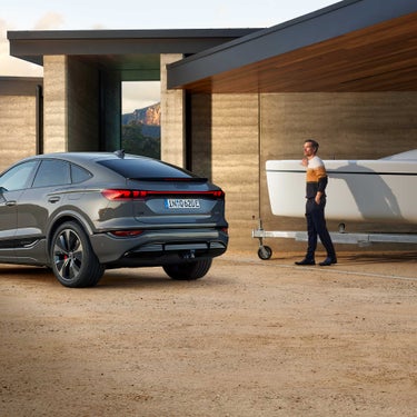 Ein grauer Audi Q6 Sportback e-tron steht vor moderner Architektur, daneben ein Mann bei einem Trailer-Boot.