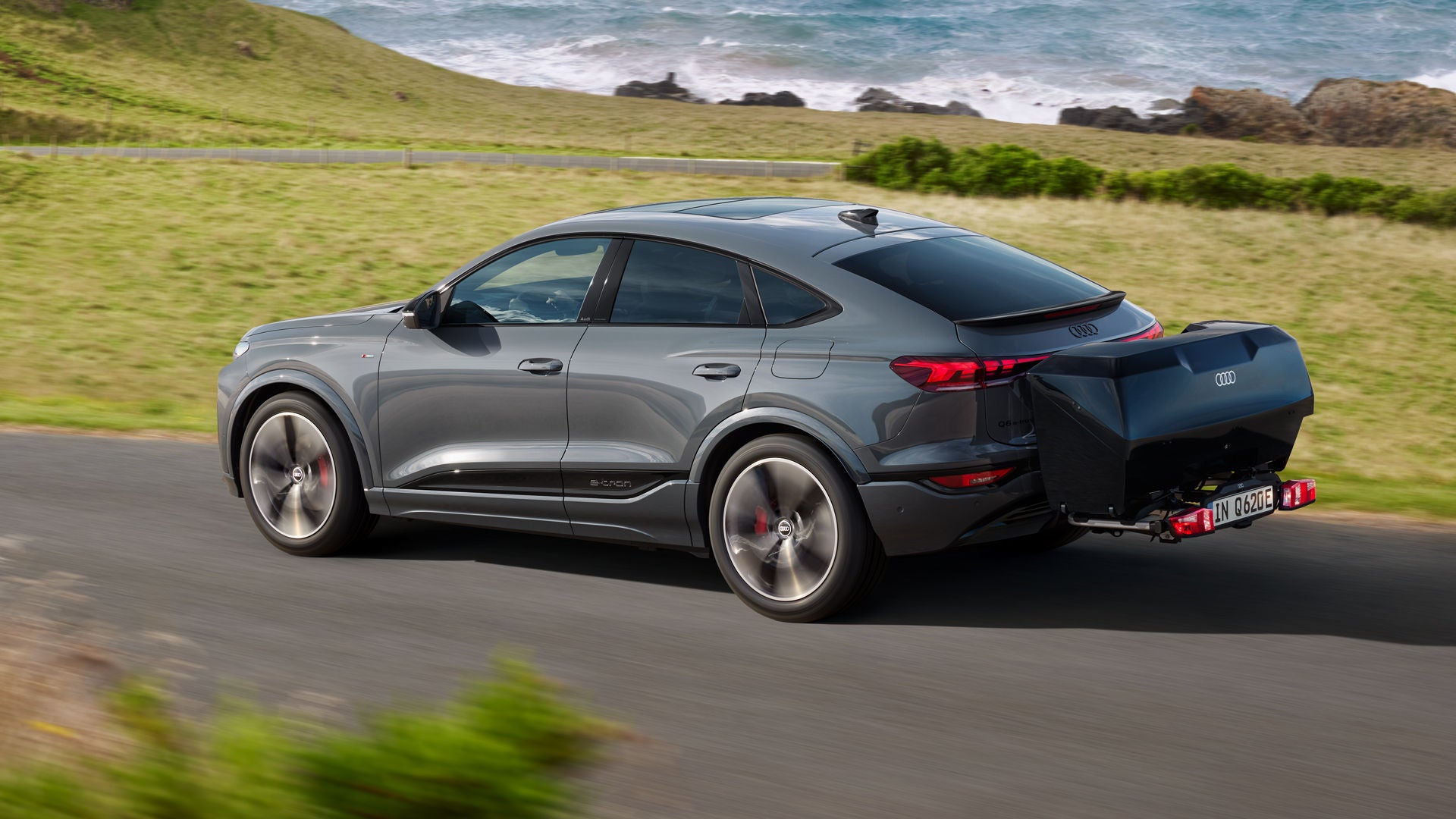 Der Q6 Sportback e-tron mit Heckbox fährt über eine Landstraße in der Natur.