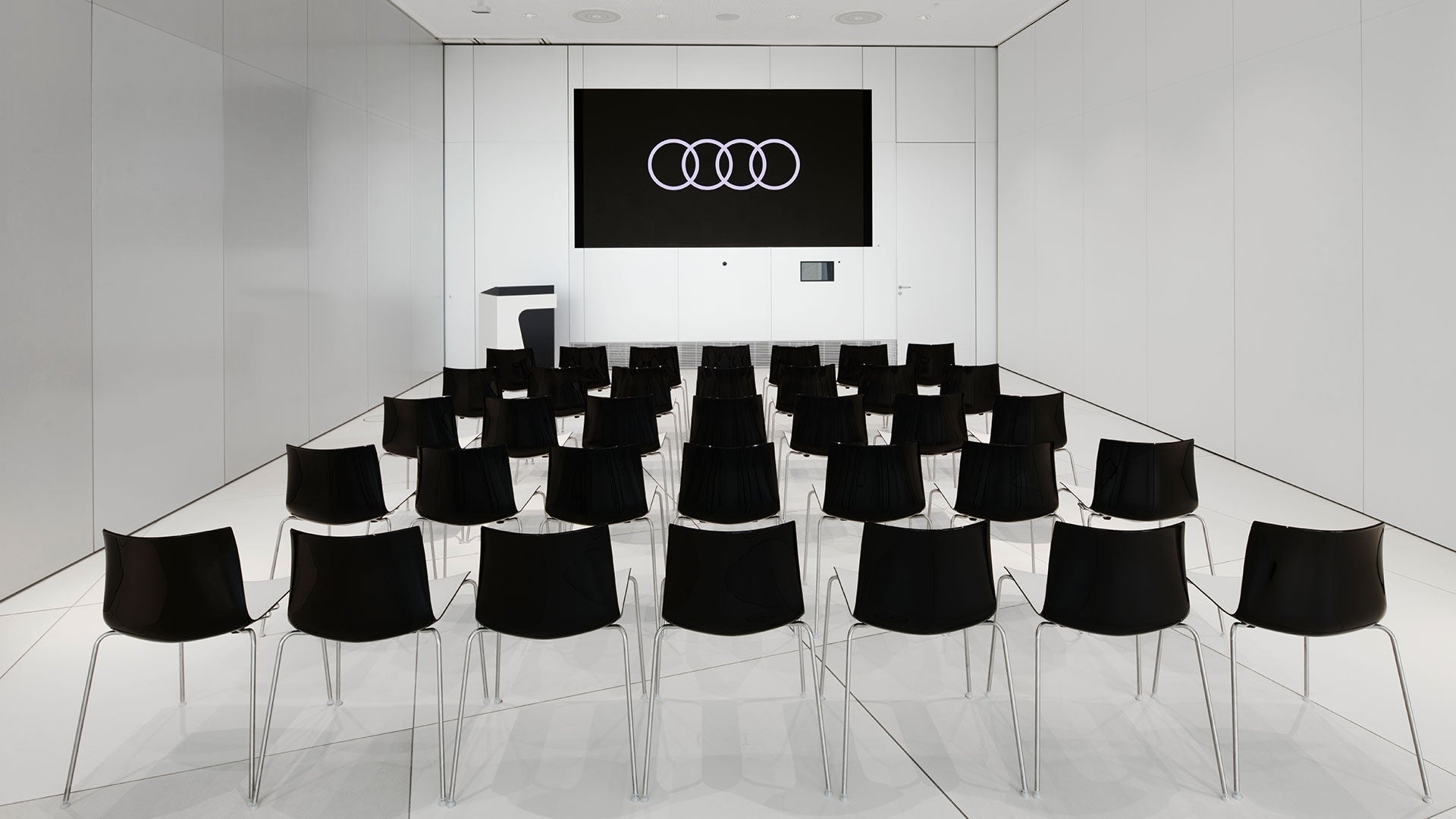 Kleiner, weißer Vortragssaal mit schwarzer Bestuhlung und Audi Logo auf großem Bildschirm.