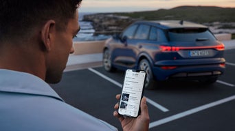 Mann blickt auf Smartphone mit Audi App, im Hintergrund parkt ein blauer Audi Q6 e-tron auf einem Küstenparkplatz.