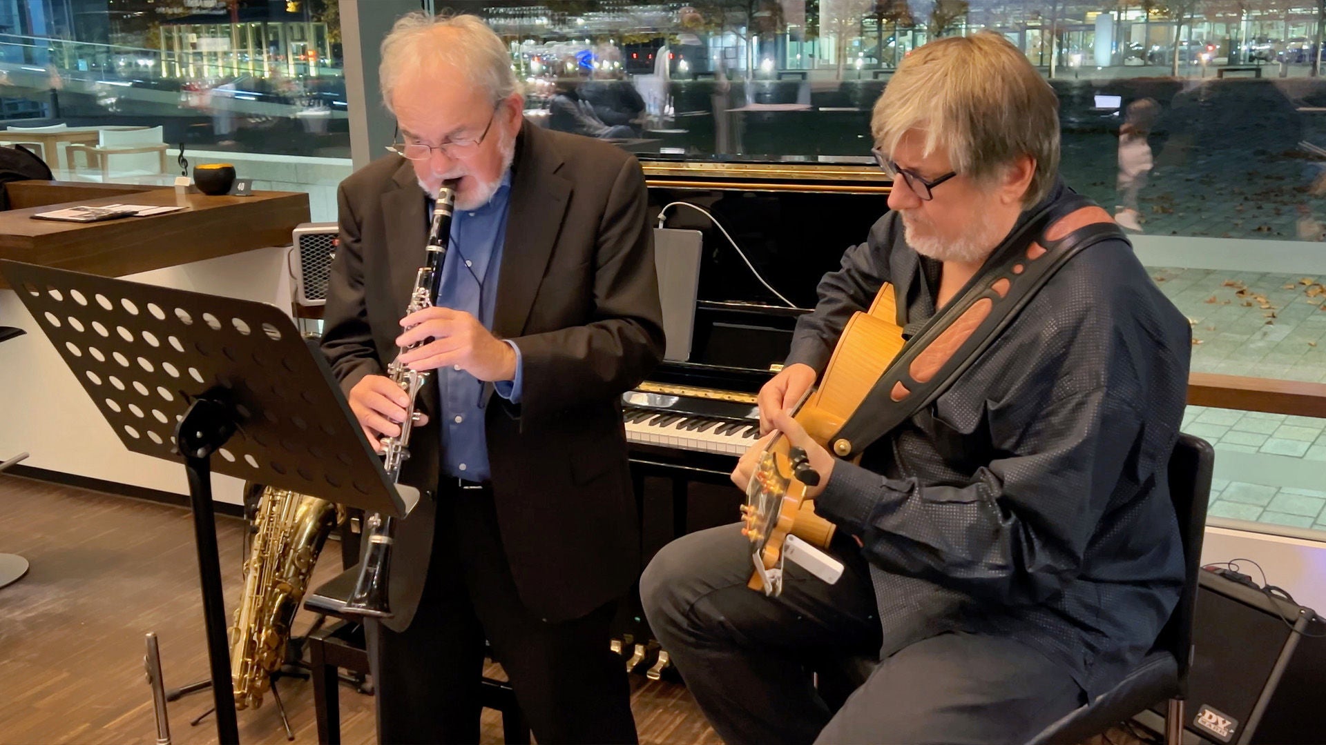 Zwei Jazzmusiker spielen Klarinette und Gitarre vor einem Klavier, Notenständer im Vordergrund, intime Atmosphäre bei Indoor-Konzert.