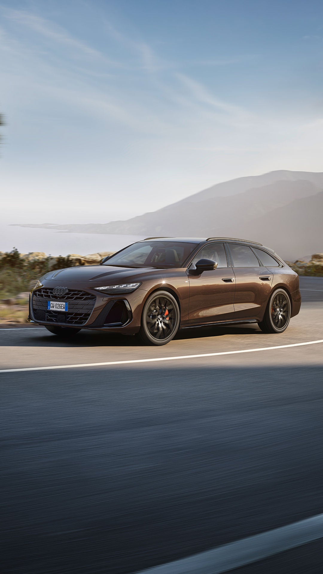 Brauner Audi A6 fährt auf kurviger Küstenstraße bei Tageslicht, Berge und Meer im Hintergrund.