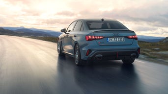 Blaue Audi RS 3 Limousine auf nasser Straße, von hinten links bei Sonnenuntergang gesehen.