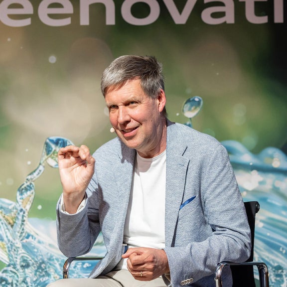 A man in a light blue jacket speaks on a stage with a microphone to his ear, the words “Greenovation” in the background.