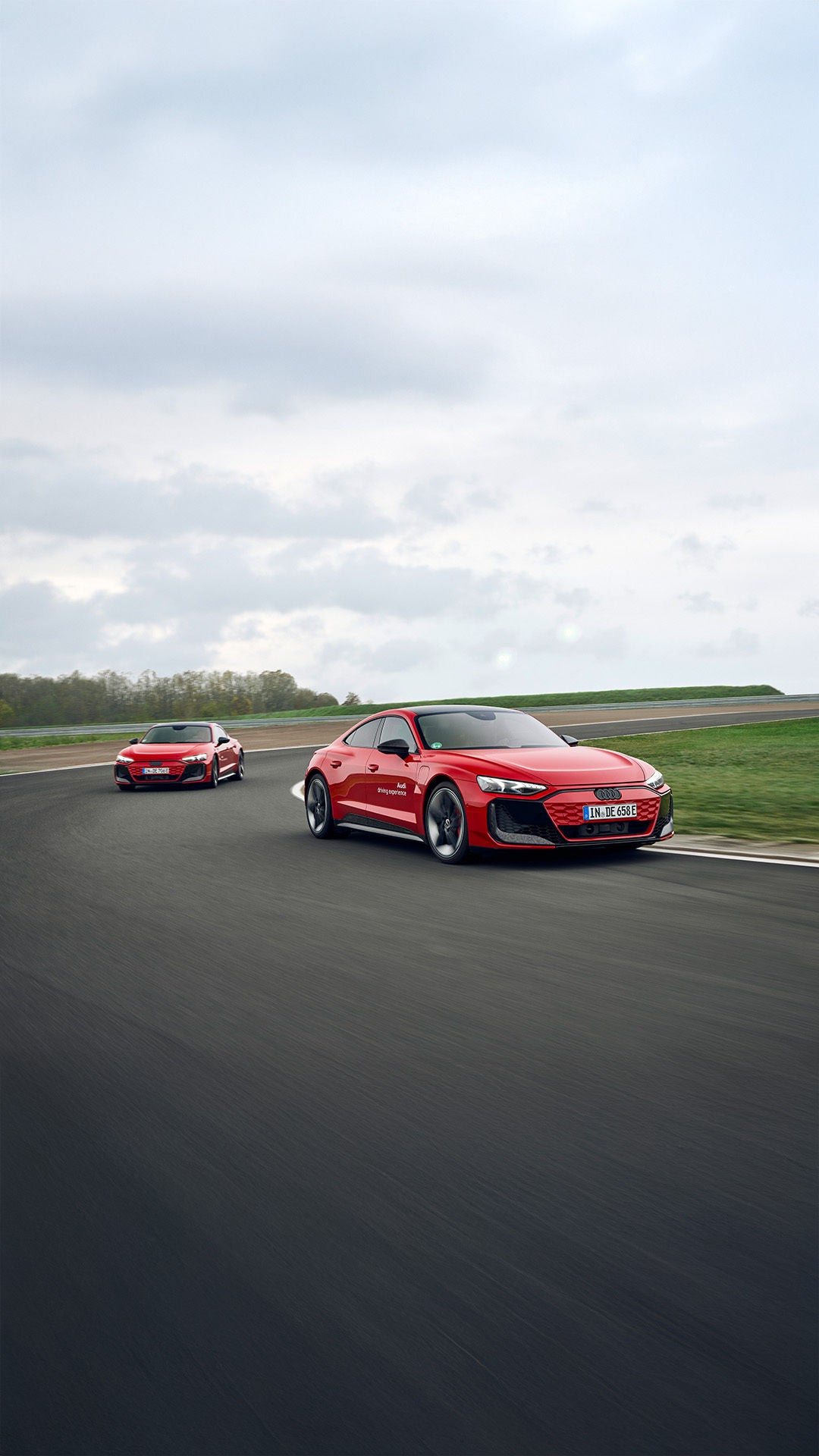 Two red Audi RS e-tron GT performance models drive one behind the other through a curve on a race track under gray skies.