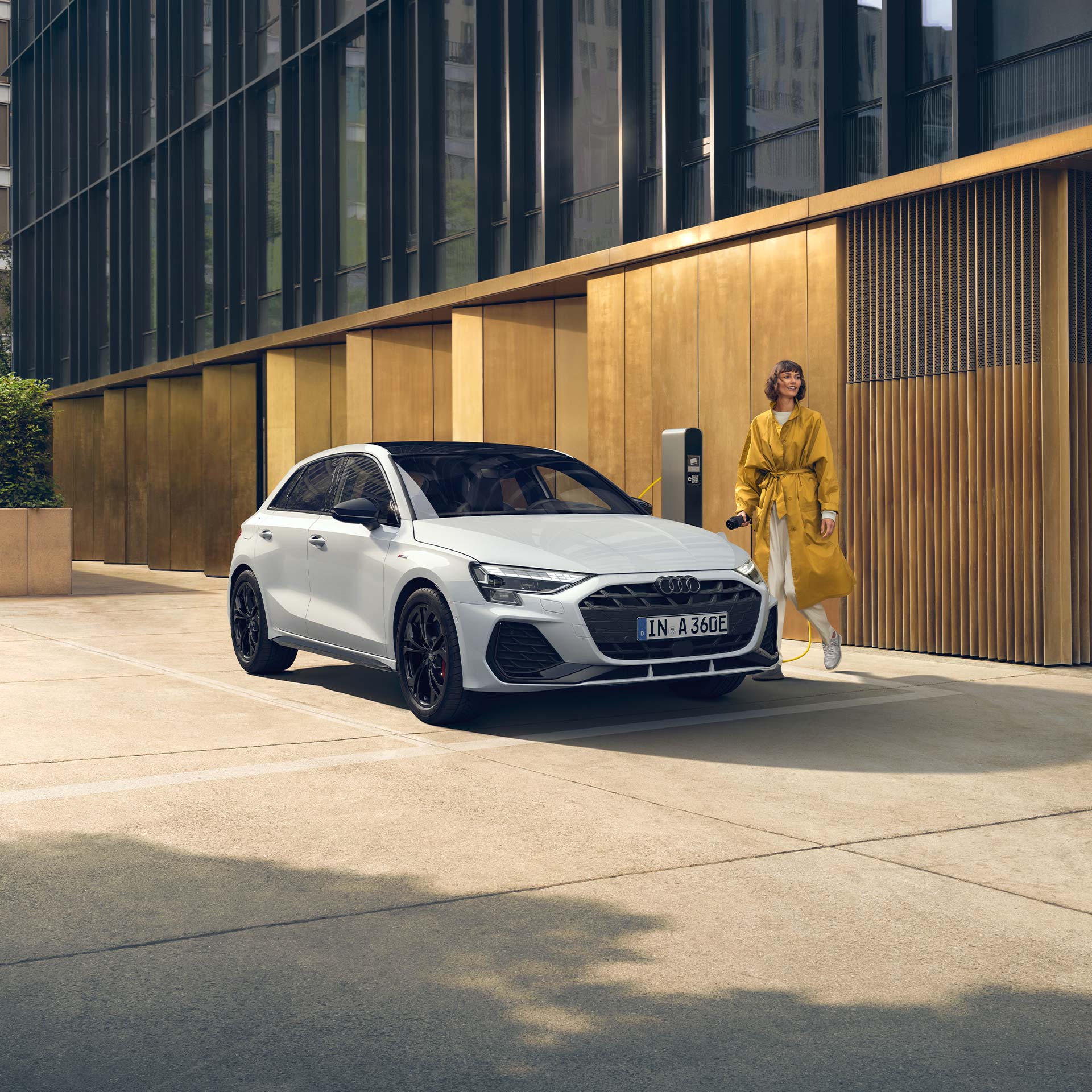Weißer Audi A3 Sportback TFSI e an Ladesäule, Frau in gelbem Mantel steht daneben vor moderner Fassade.