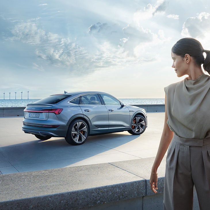 Woman looking at an Audi e-tron Sportback standing by the sea. Windmills can be seen on the horizon.
