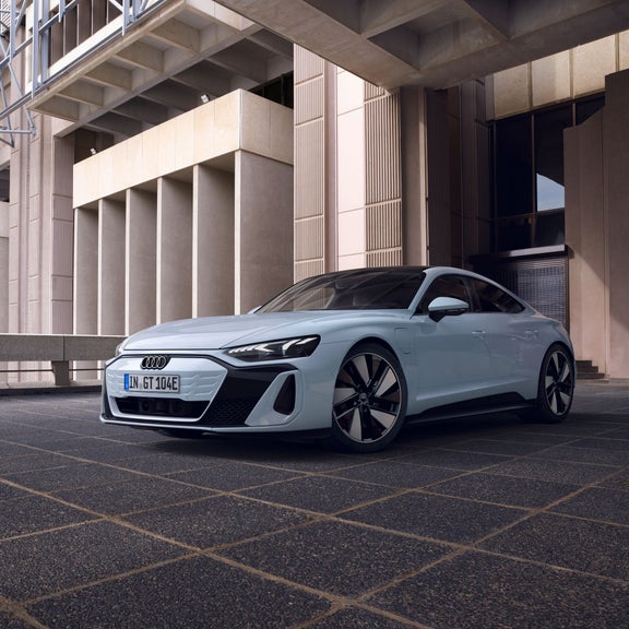 Frontansicht eines hellblauen Audi e-tron GT quattro auf dem Parkdeck eines Parkhauses mit moderner Architektur. 