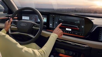 Fahrerin im gelben Outfit bedient großes Touchdisplay im Audi, Blick auf Digitalanzeigen und Landschaft bei Sonnenuntergang.