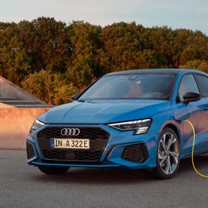 A blue Audi car is parked on a rooftop, charging with a yellow cable. The background features lush green trees under a clear sky at sunset.