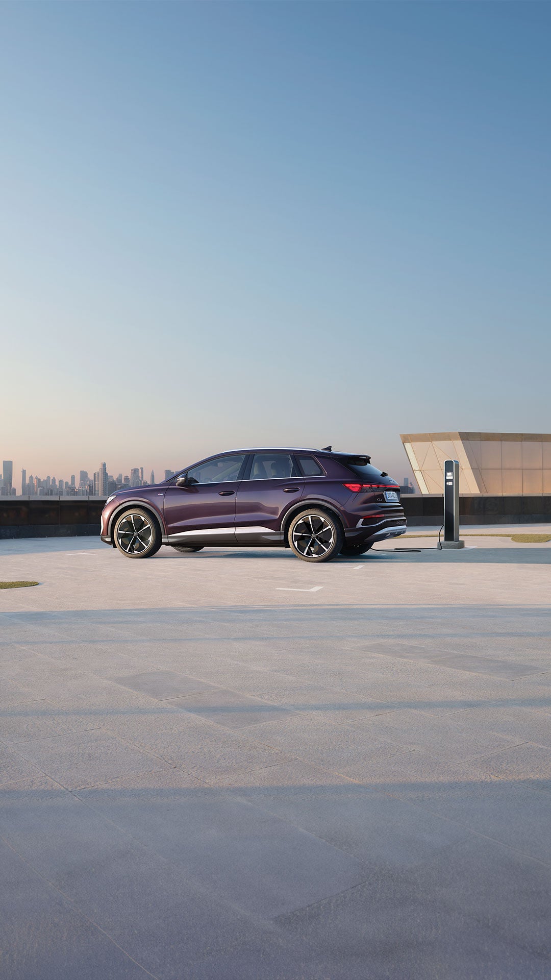 Seitenansicht eines violetten Audi Q4 e-tron mit sportlichen Felgen an einer Ladesäule auf Parkdeck mit Stadtpanorama im Hintergrund.