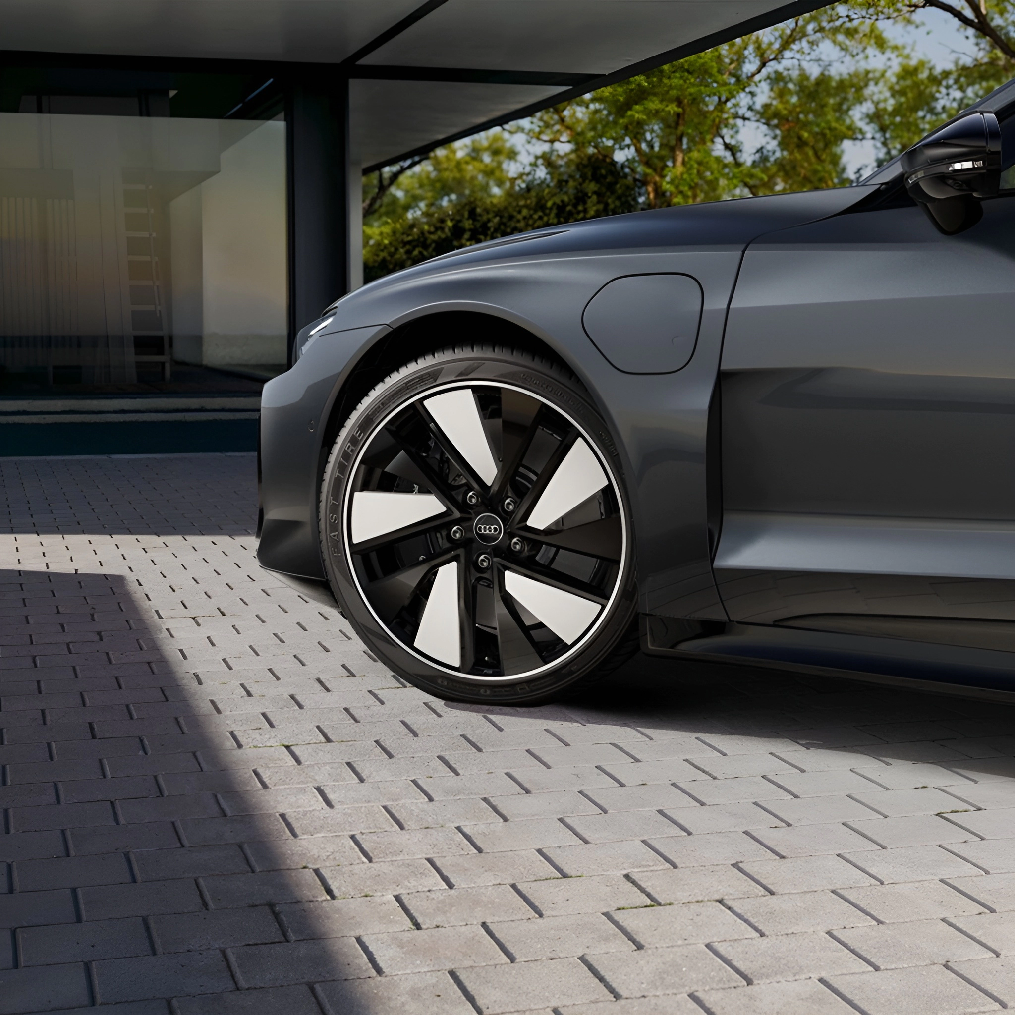 Grey Audi car on brick driveway, showing front wheel and part of body, with trees and building in background.