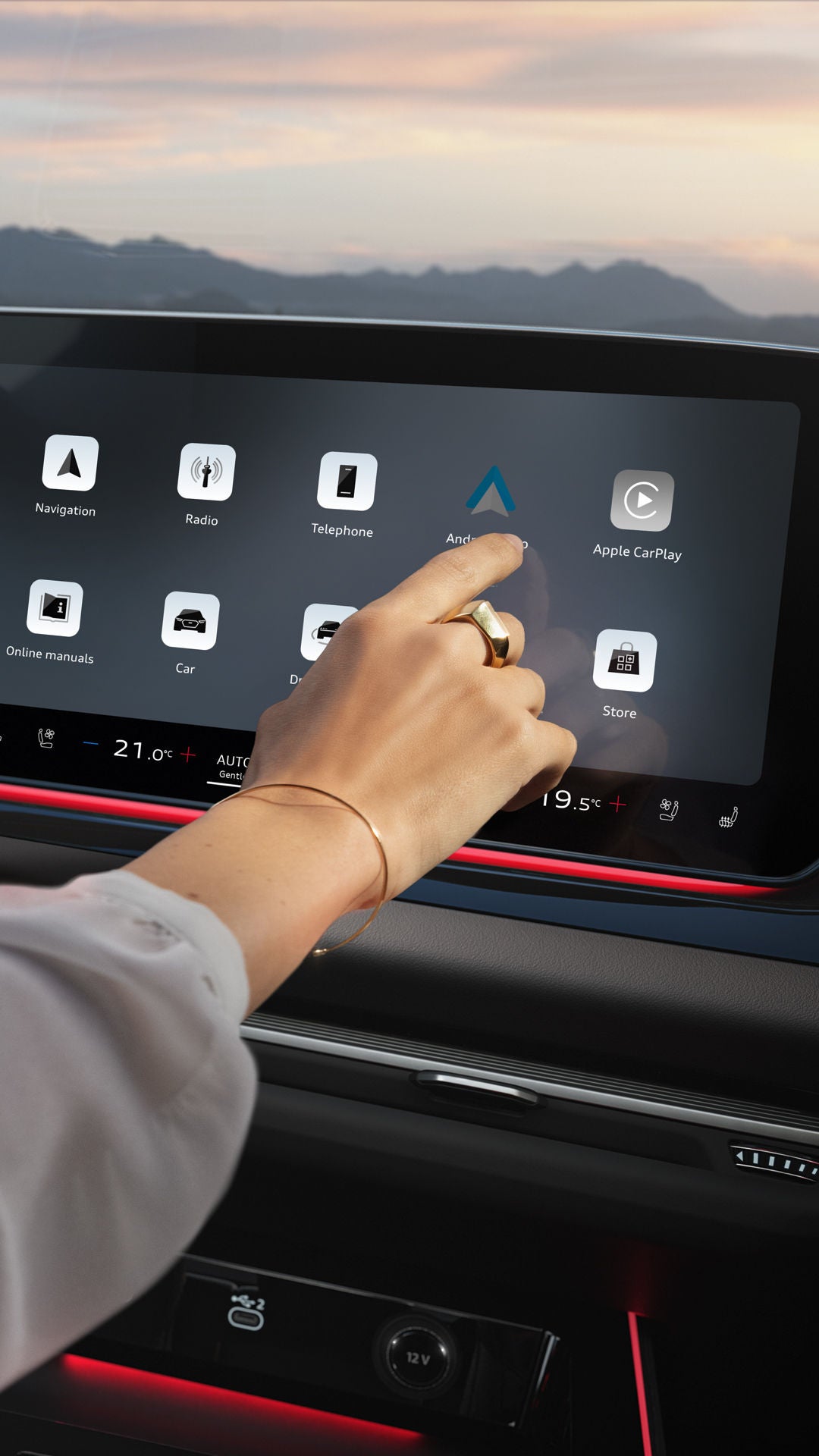 Close-up van de cockpit van een Audi: het stuur aan de linkerkant, een verlicht display met app-pictogrammen in het midden.  De arm van een vrouw strekt zich vanaf de linkerkant in het beeld uit en tikt op het display.