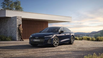 Ein blauer Audi vor modernem Haus; Person lehnt an Steinwand, Berge und Abendhimmel im Hintergrund.