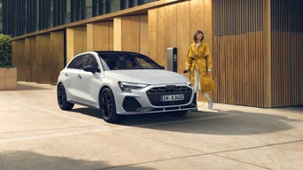 Weißer Audi A3 Sportback TFSI e steht vor einer goldenen Fassade, daneben eine Frau in gelbem Mantel an der Ladestation.