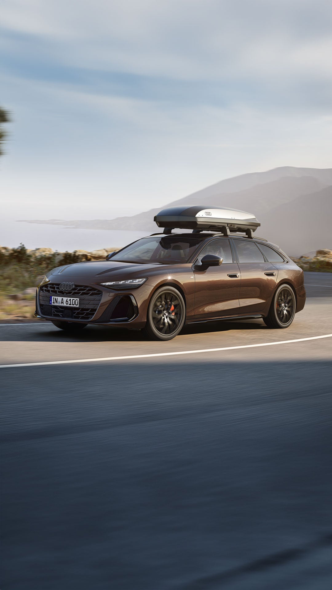 Brauner Audi-Kombi mit Dachbox fährt auf Küstenstraße entlang Felsenlandschaft und Bergen bei leichtem Nebel im Hintergrund.