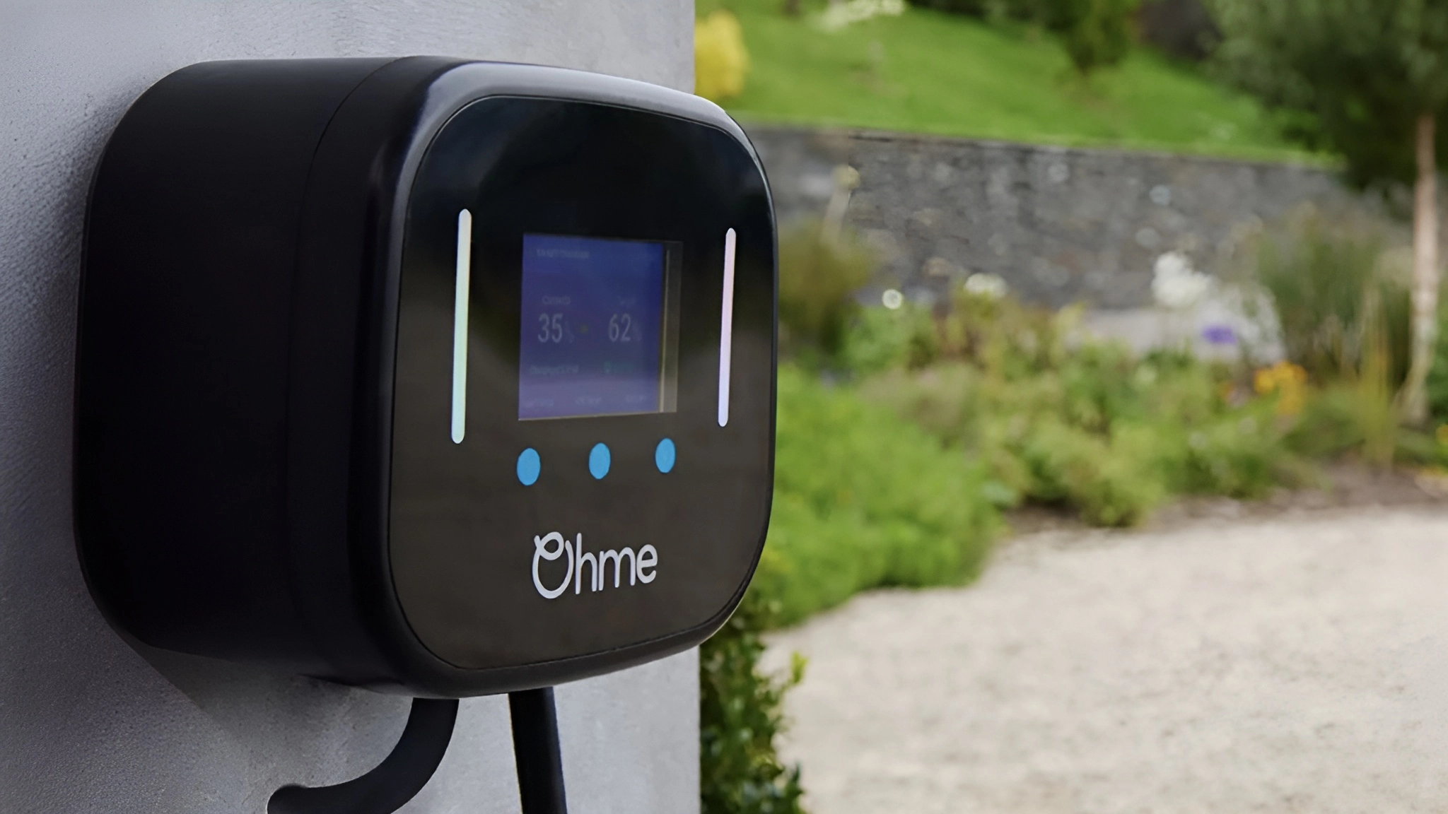 Black Ohme charger on wall with screen displaying 35% and 62, blue buttons, and white logo; background shows blurred garden and stone wall.