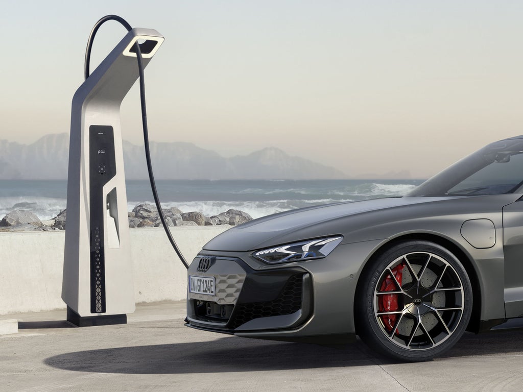 Ein grauer Audi RS e-tron GT lädt an einer Ladestation an der Küste mit Wellen und Bergen im Hintergrund.