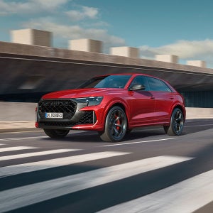 Red Audi with a modern design drives across a zebra crossing on an urban road. The background features a concrete structure under a clear sky.
