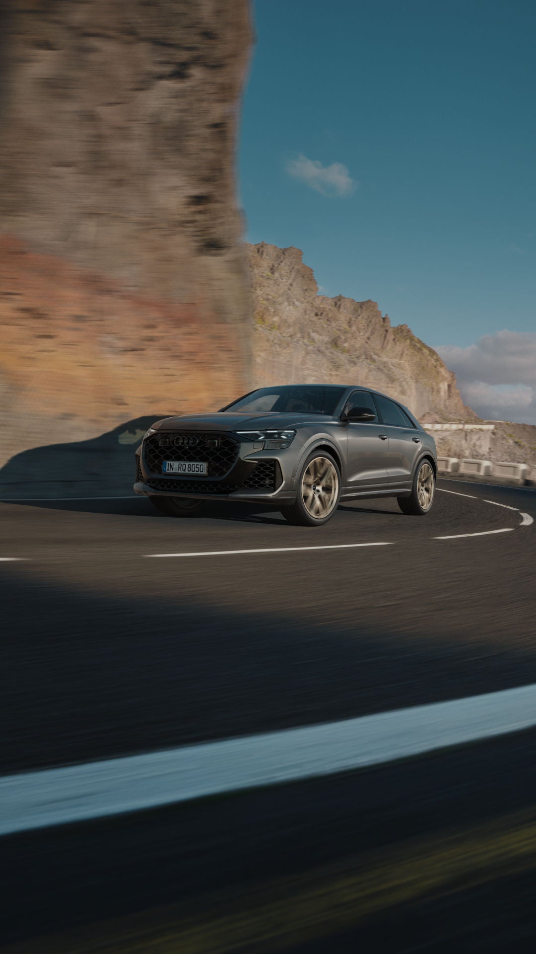 A sleek grey Audi cruises around a coastal bend, with rocky cliffs and a blue sky creating a dramatic backdrop.