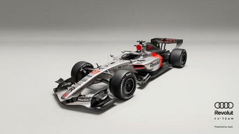 Schrägansicht eines silber-schwarzen Formel-1-Rennwagens mit roten Akzenten vor grauem Hintergrund, Audi- und Revolut-Logo in schwarz am Rand.