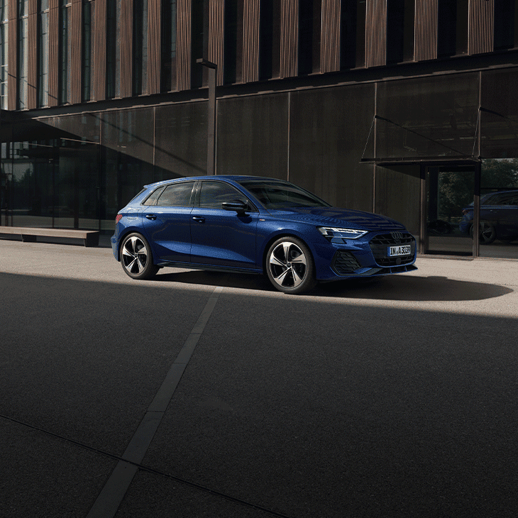 Une A3 bleue foncée est stationnée sur une route asphaltée devant un bâtiment moderne aux façades vitrées sombres. La scène est éclairée par une lumière naturelle créant des reflets sur la carrosserie et de longues ombres sur le sol.