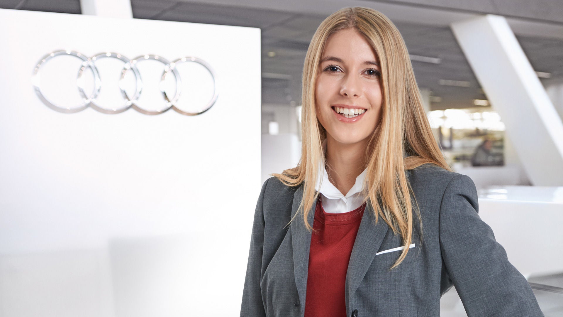 Eine lächelnde junge Frau in einem grauen Blazer steht an einem Verkaufstresen mit den Audi Ringen auf einer Trennwand hinter ihr. 