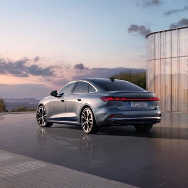 Graue Audi A5 Limousine steht vor Glasfassade bei Sonnenuntergang, Berge und Himmel im Hintergrund.