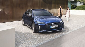 Dunkelblauer Audi RS 6 Avant parkt auf Kopfsteinpflaster, im Hintergrund ein Paar vor moderner Fassade.