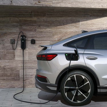 Silberner Audi Q4 Sportback e-tron mit sportlichen Felgen wird an einer Wandladestation geladen, seitliche Heckansicht vor Steinwand.