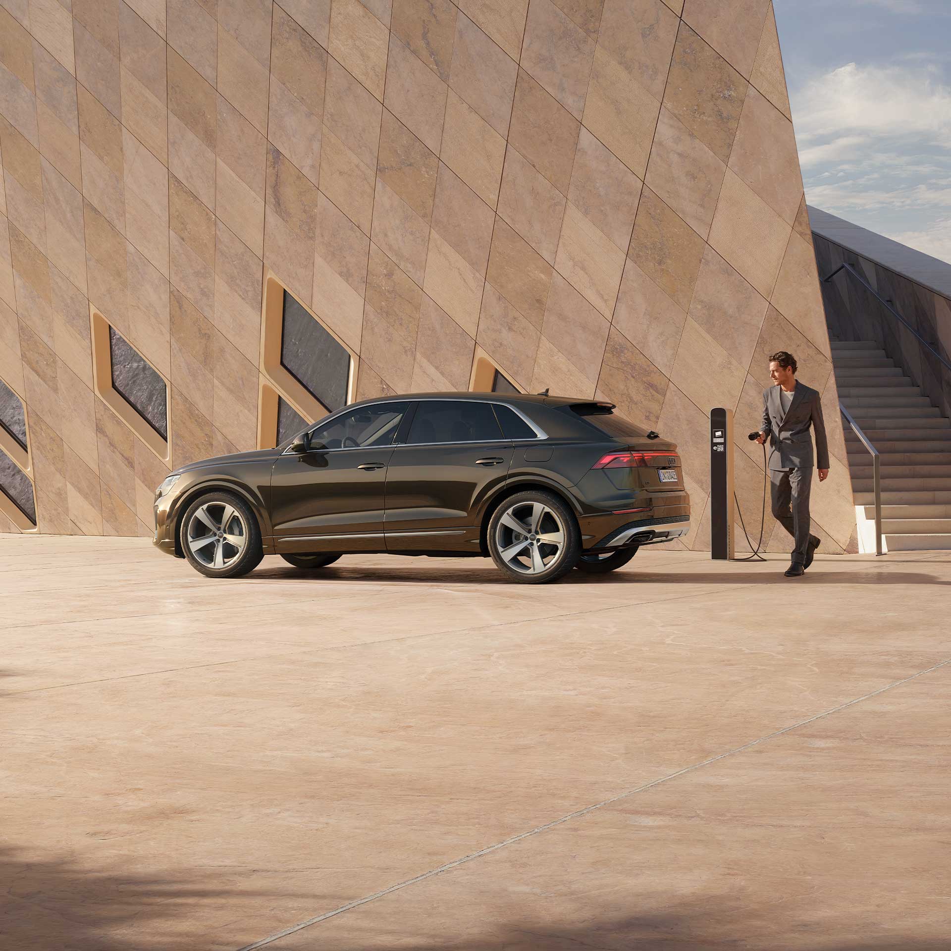 Ein Mann mit Ladekabel steht neben einem braunen Audi Q8 SUV TFSI e vor einem modernen Gebäude mit diagonalen Fenstern.