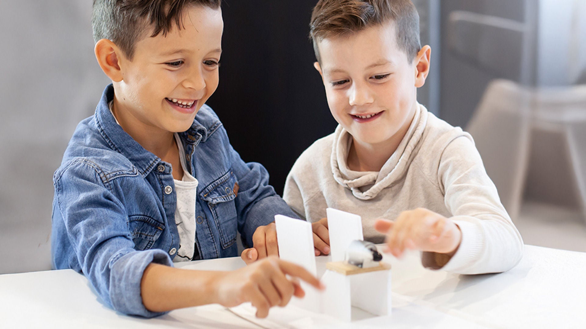 zwei Kinder sitzen gemeinsam an einem Tisch und experimentieren wie sie ein Fahrzeug mit Hilfe eines Luftkissens fortbewegen können