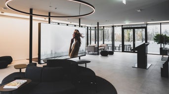 Moderner Loungebereich mit Sofas, großem Display mit Frau im Wind und Glasfront mit Ausblick auf Bäume.