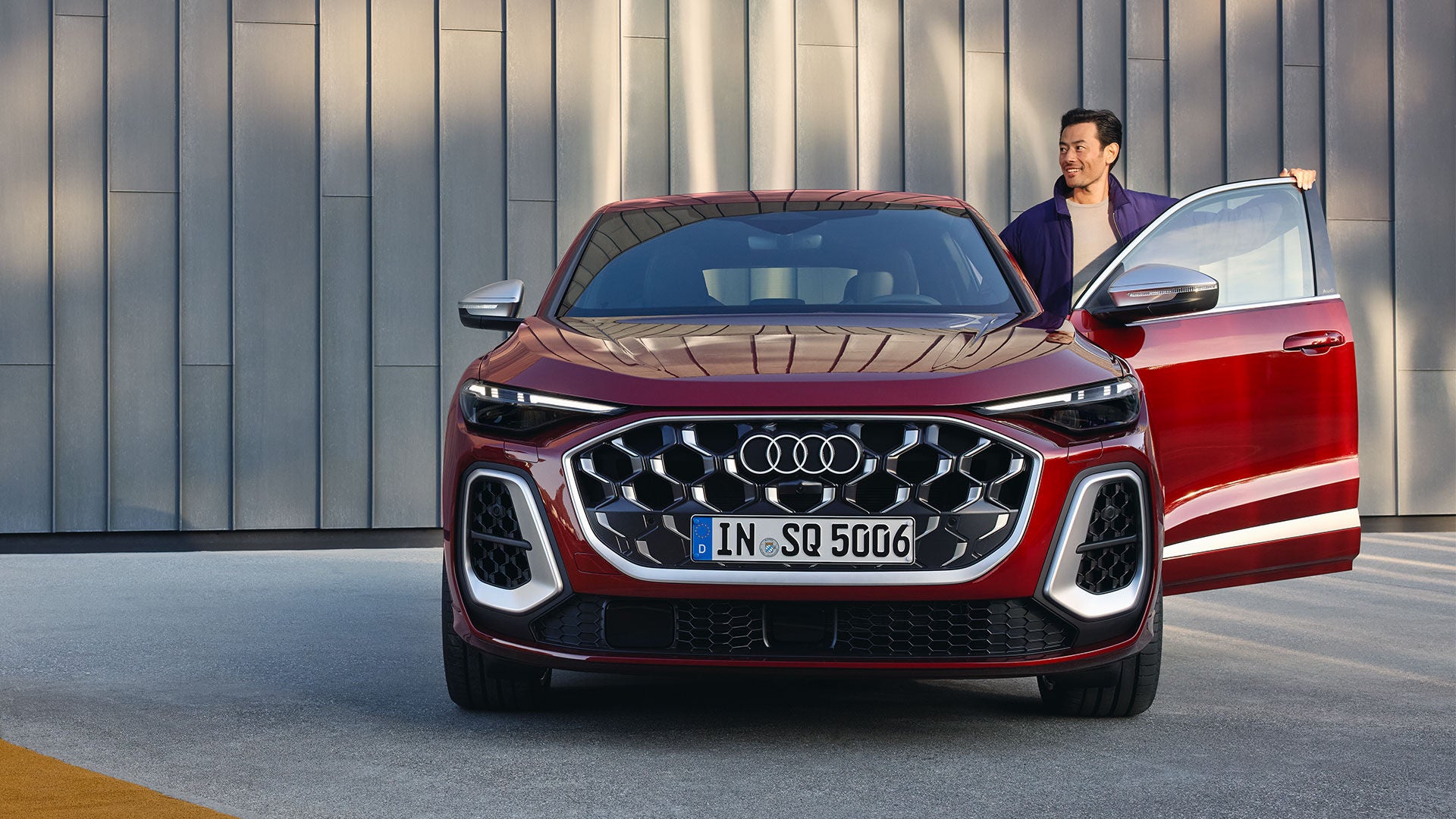 Un homme aux cheveux foncés se tient dans la portière ouverte d'une Audi SQ5 rouge, que l'on voit de face.