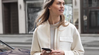 Frau mit Smartphone steht draußen vor einem geparkten Auto und schaut zur Seite.