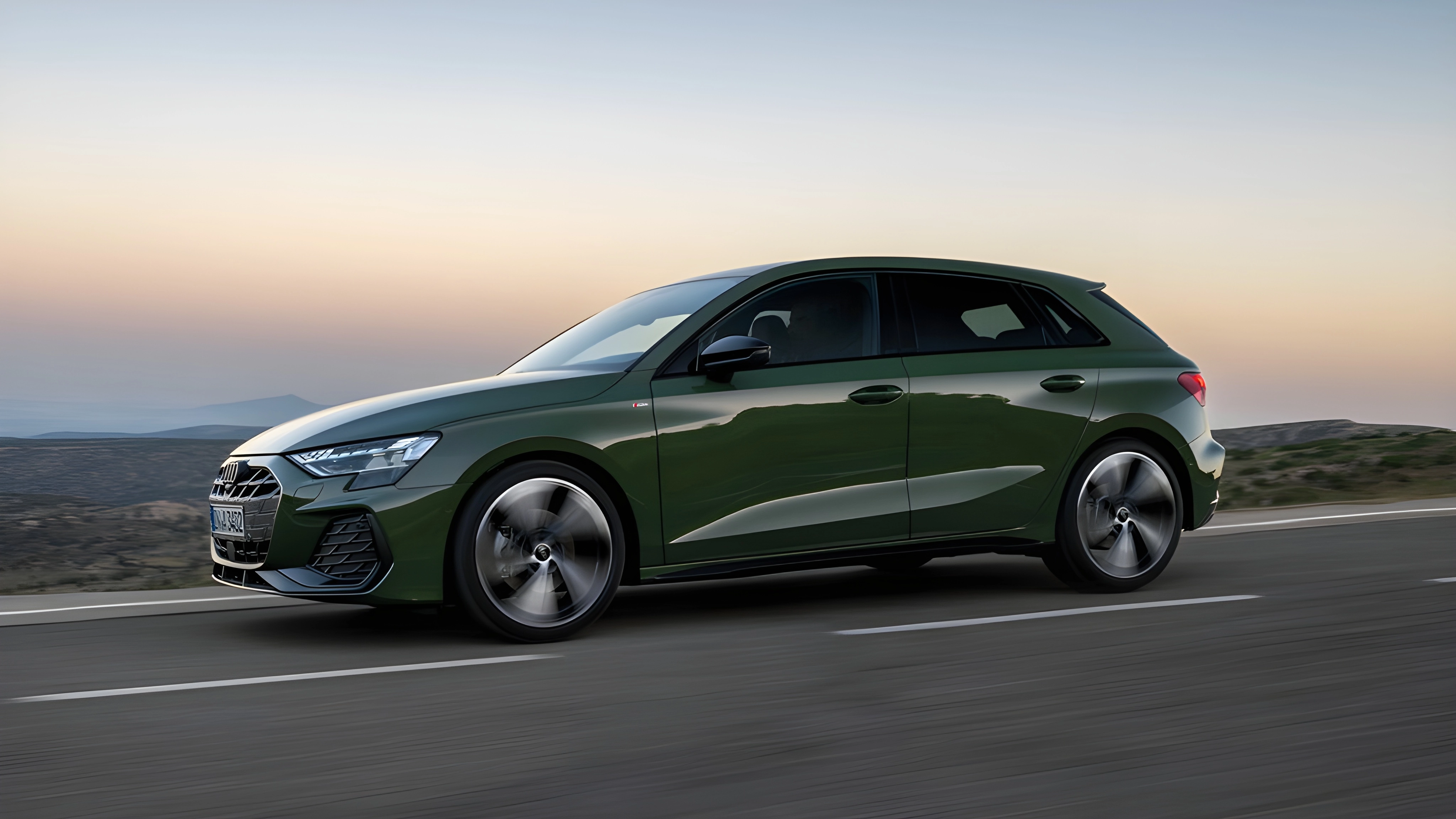 Green Audi A3 Sportback driving on a road, set against a backdrop of hills and a clear gradient sky.