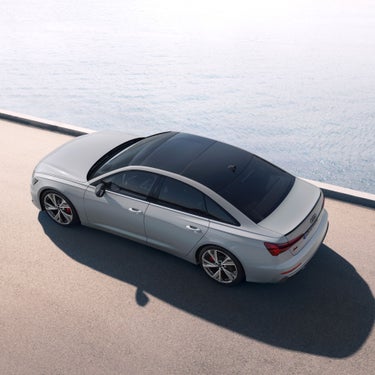 A silver sedan parked beside calm water, viewed from above with clear skies.