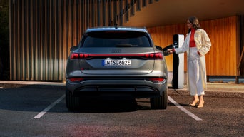 Der SQ6 SUV e-tron steht auf einem Parkplatz vor einer Gebäudefassade und wird von einer jungen Frau an eine Ladestation angeschlossen.