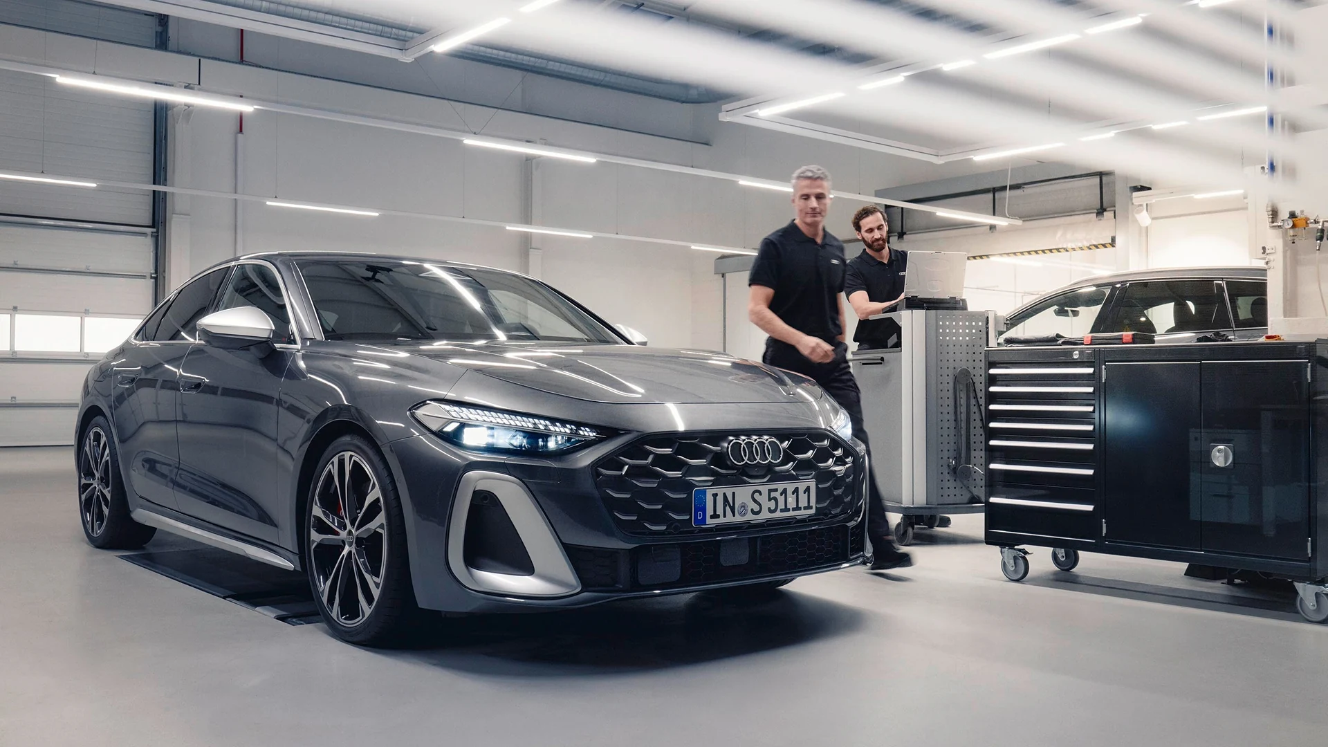 Audi Sportback e-tron undergoing service inspection in a modern workshop, shown from the front three-quarter angle. The vehicle features LED headlights, a sculpted front grille and large alloy wheels. In the background, technicians work at a diagnostic workstation beside tool cabinets under bright overhead lighting, highlighting the professional service environment.